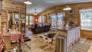 L’intérieur d’un chalet rustique comprend un coin repas, un canapé, un comptoir de cuisine et une grande fenêtre agrémentée de rideaux.