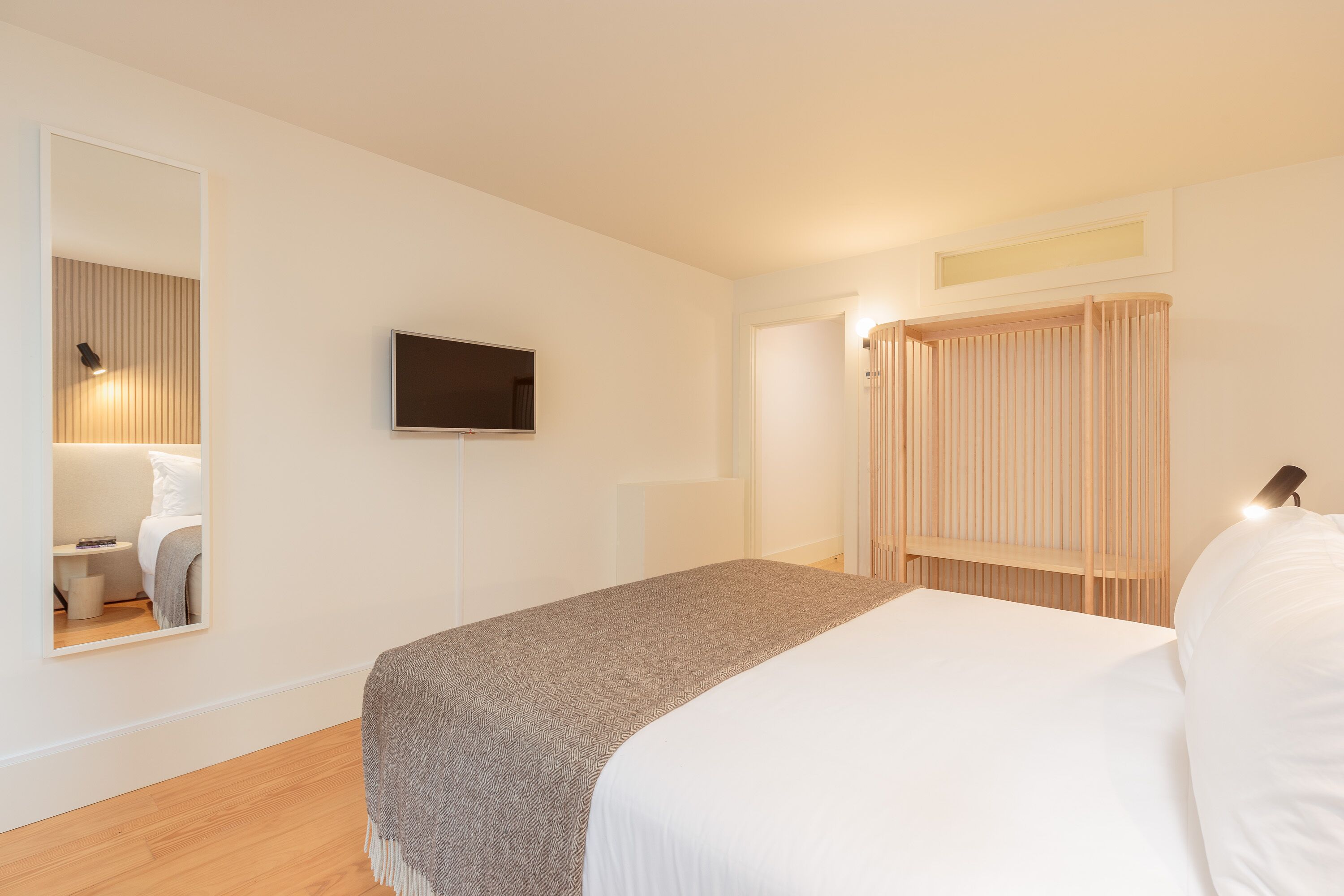 A modern hotel room with a bed, a TV, a mirror, and a wooden wardrobe.