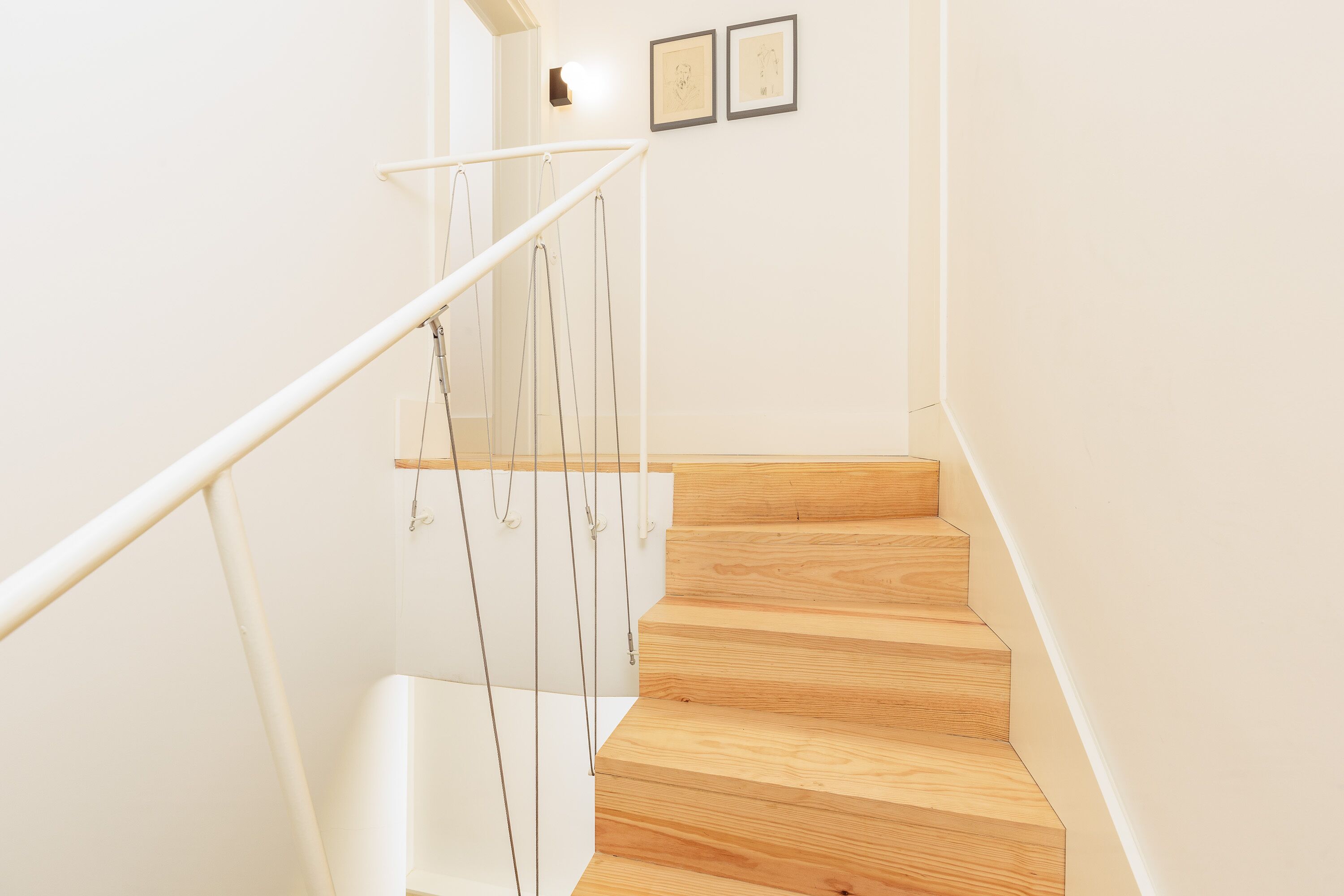 A wooden staircase with a glass railing, white walls, and two framed pictures on the wall.