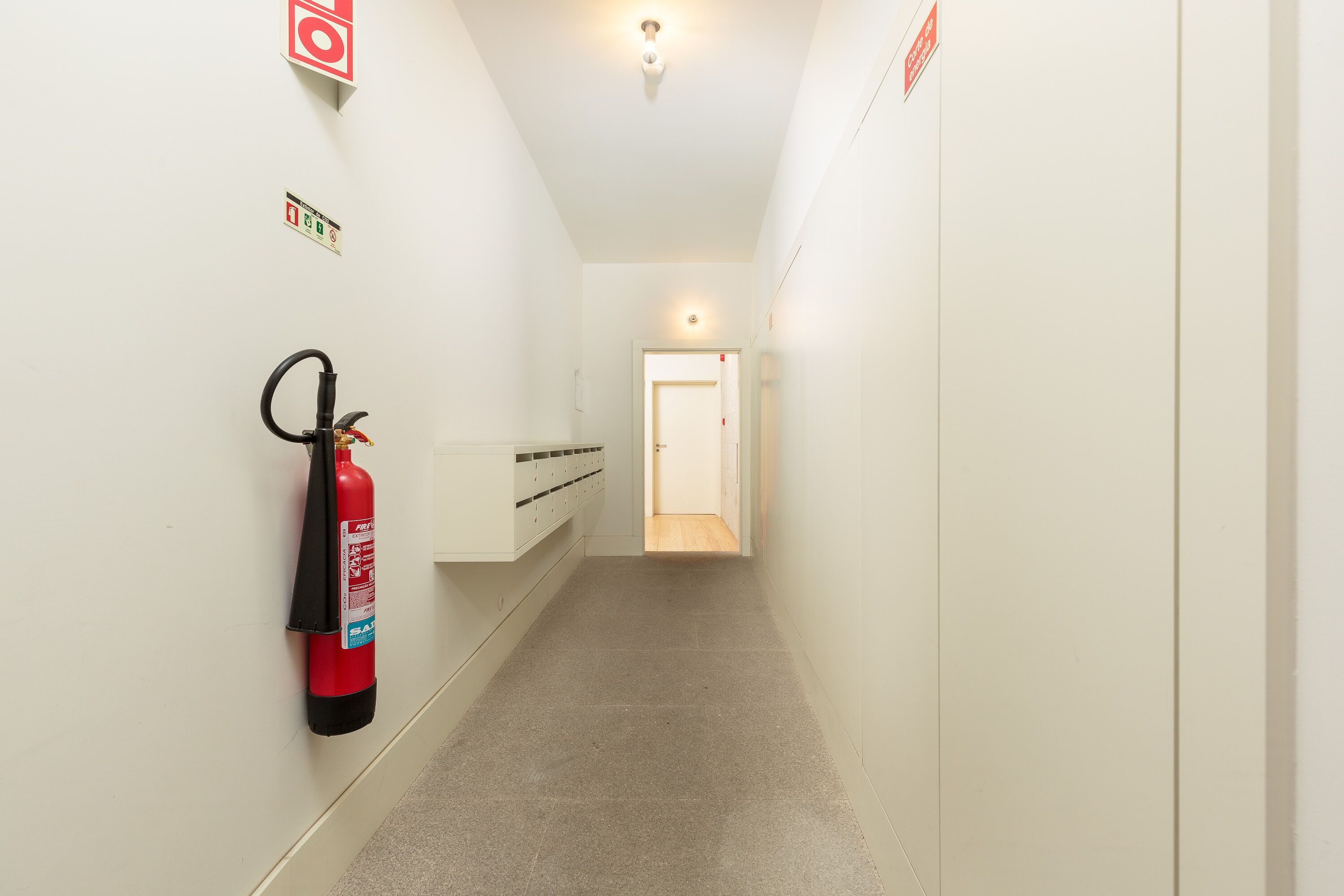 A narrow hallway with a fire extinguisher mounted on the wall, a light fixture on the ceiling, and a door at the end.