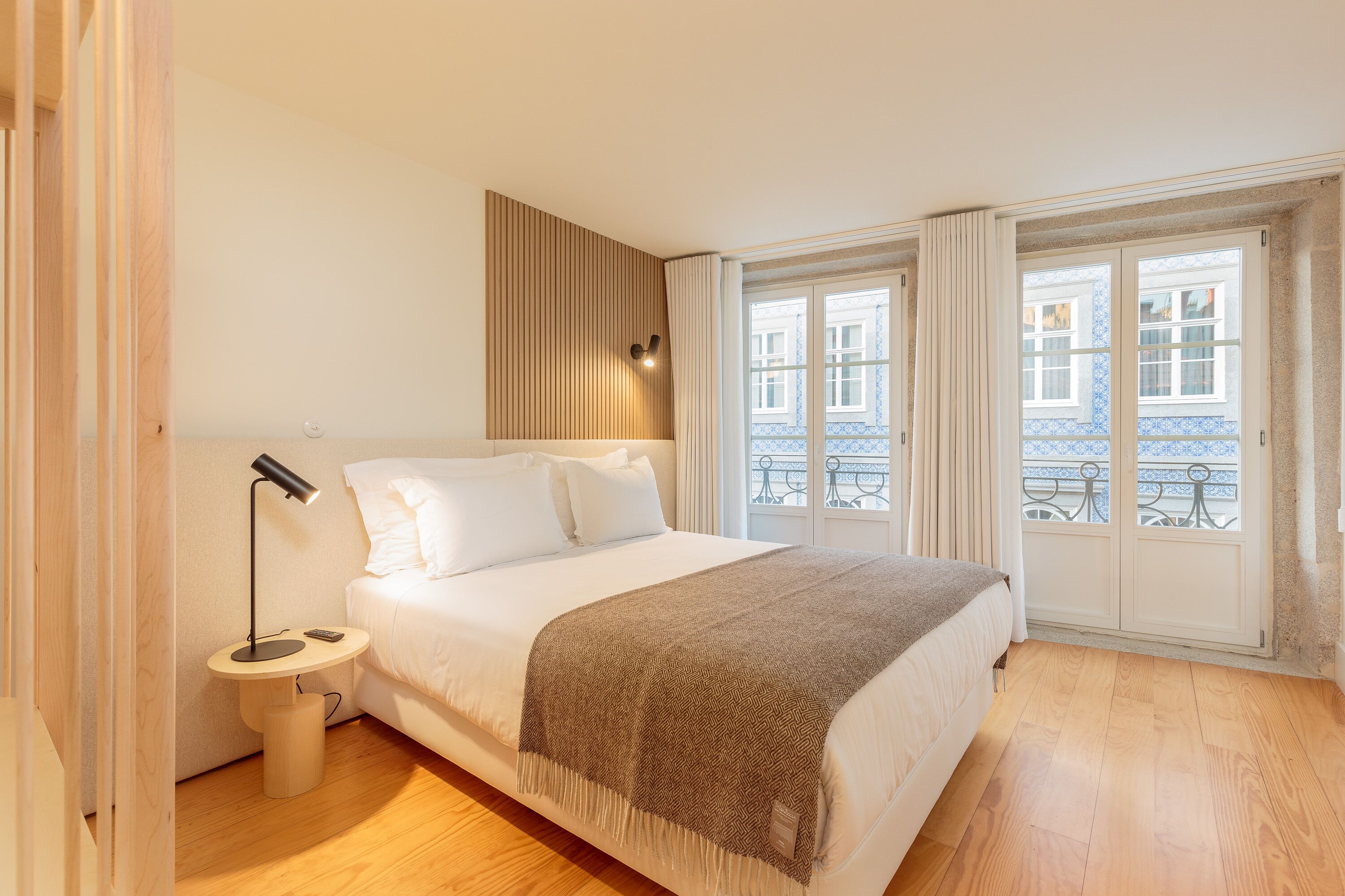 A bedroom with a bed, bedside table, lamp, and a view of buildings through a window.