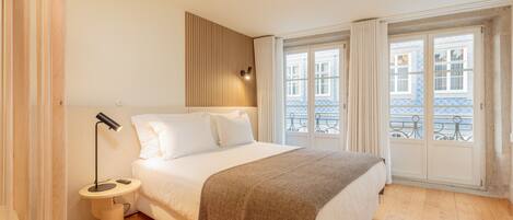 A bedroom with a bed, bedside table, lamp, and a view of buildings through a window.