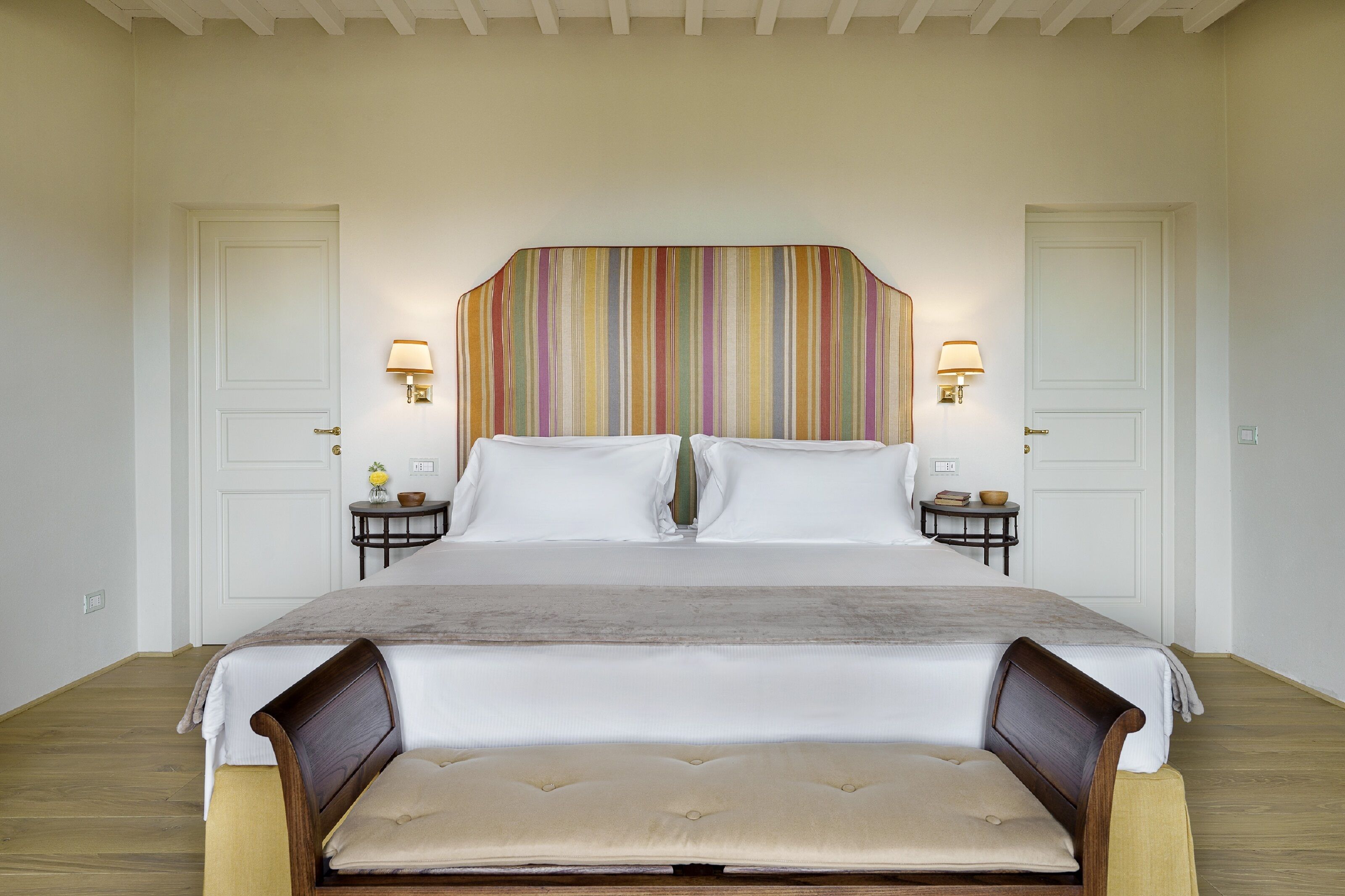 Una cama grande con cabecera de madera, dos mesitas de noche con lámparas, y un banco al pie de la cama.