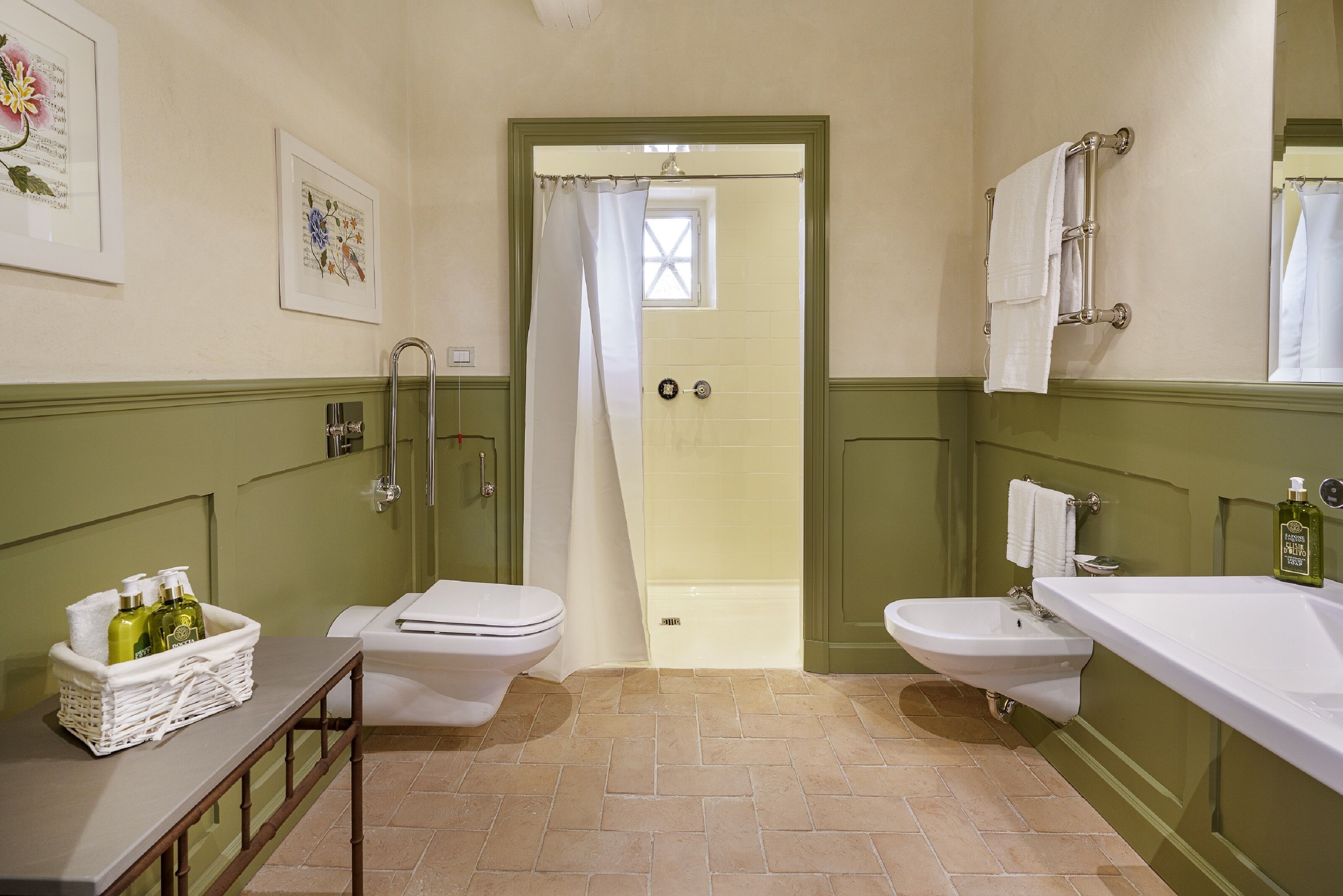 A bathroom with two toilets, a sink, a shower, and a bathtub. There are framed pictures on the wall and a basket with toiletries on the counter.