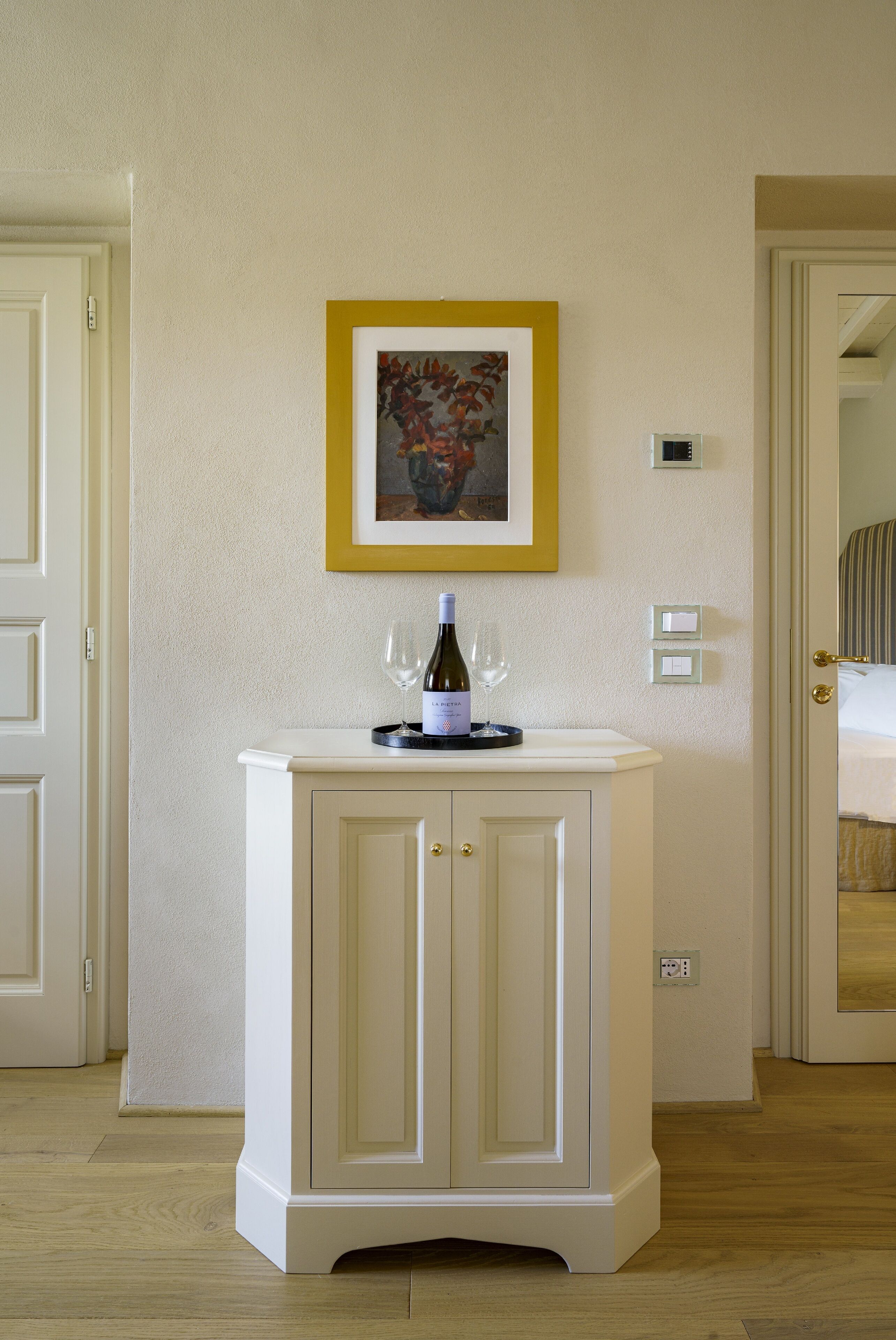 A white cabinet with a framed painting, a bottle of wine, and two glasses on top.