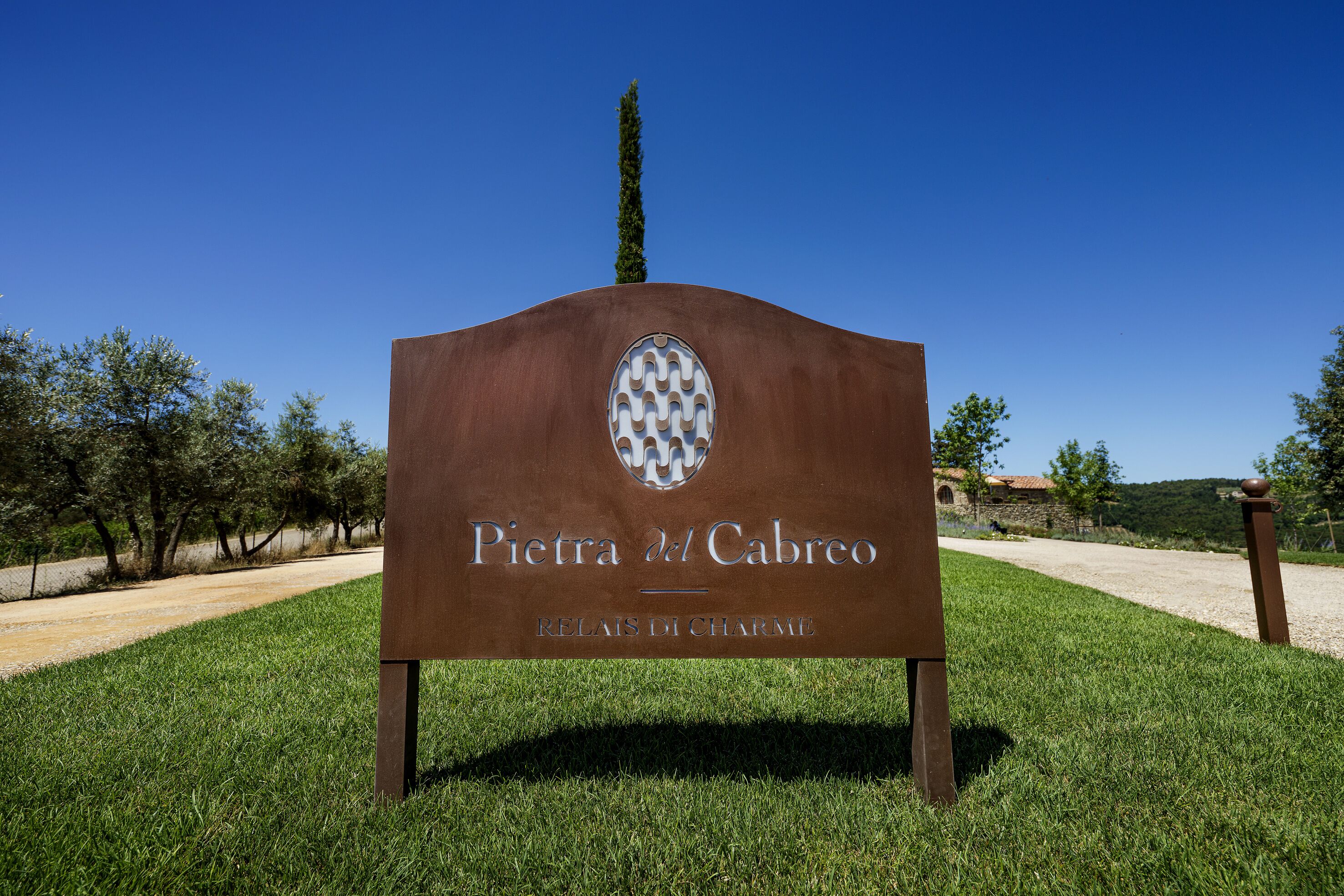 Een bord met de tekst 'Pietra del Cabreo' en een logo, gelegen in een grasrijk gebied met bomen en een heldere lucht.