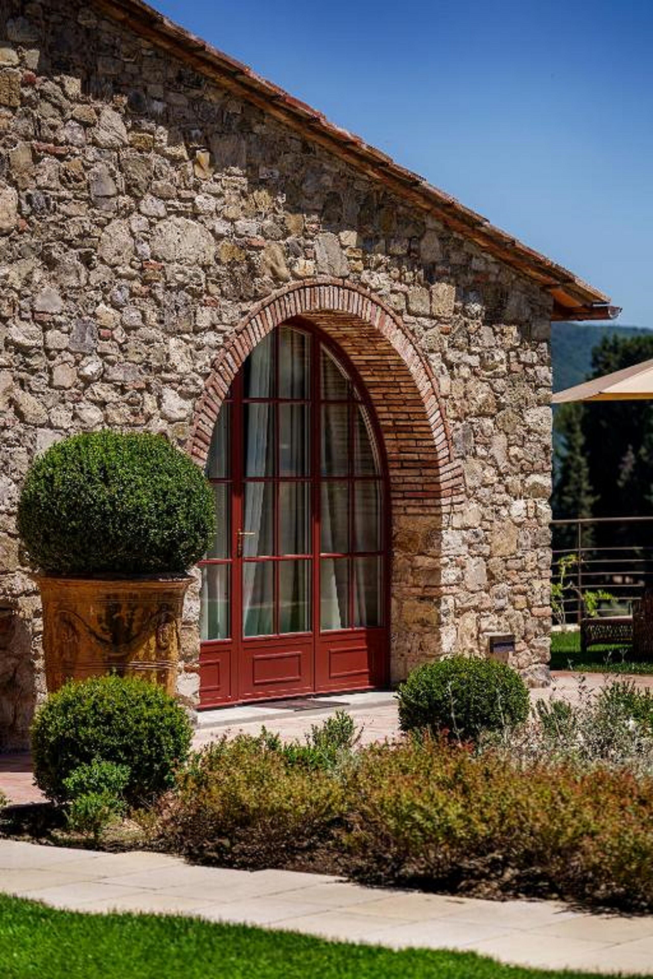 A stone building with a red door and arched window, surrounded by greenery.