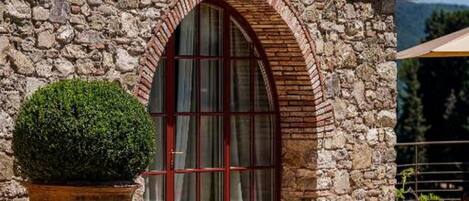 A stone building with a red door and arched window, surrounded by greenery.