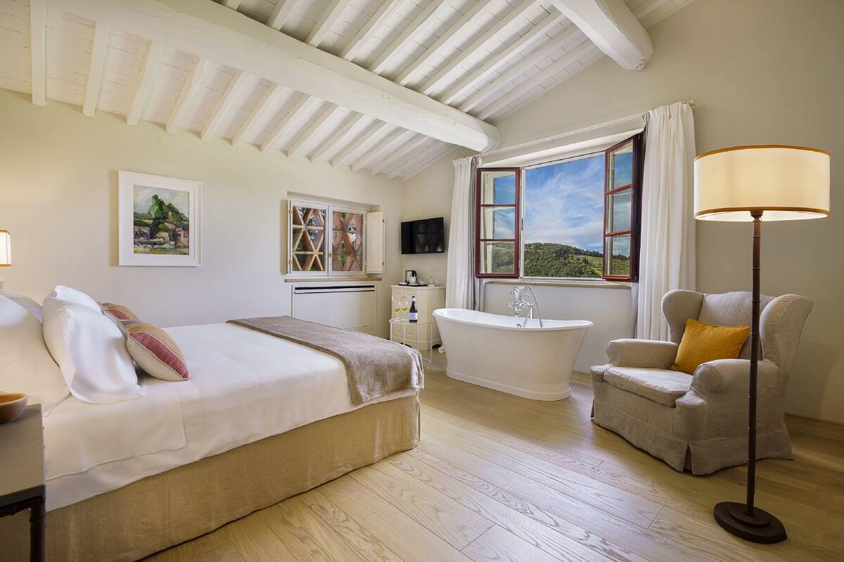 A bedroom with a bed, a bathtub, a chair, and a window with a view.