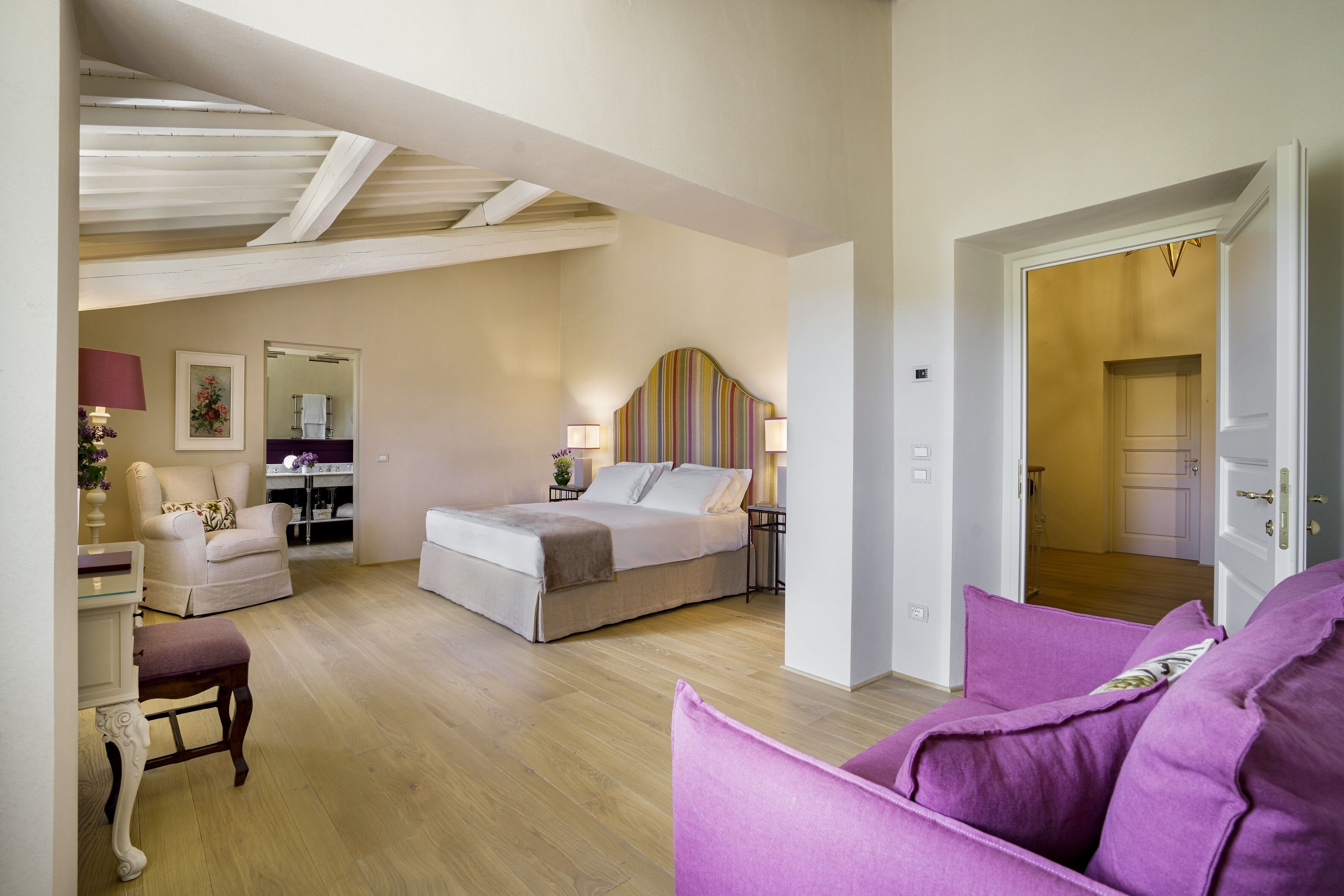 A bedroom with a bed, bedside table, lamp, chair, and a view into another room.