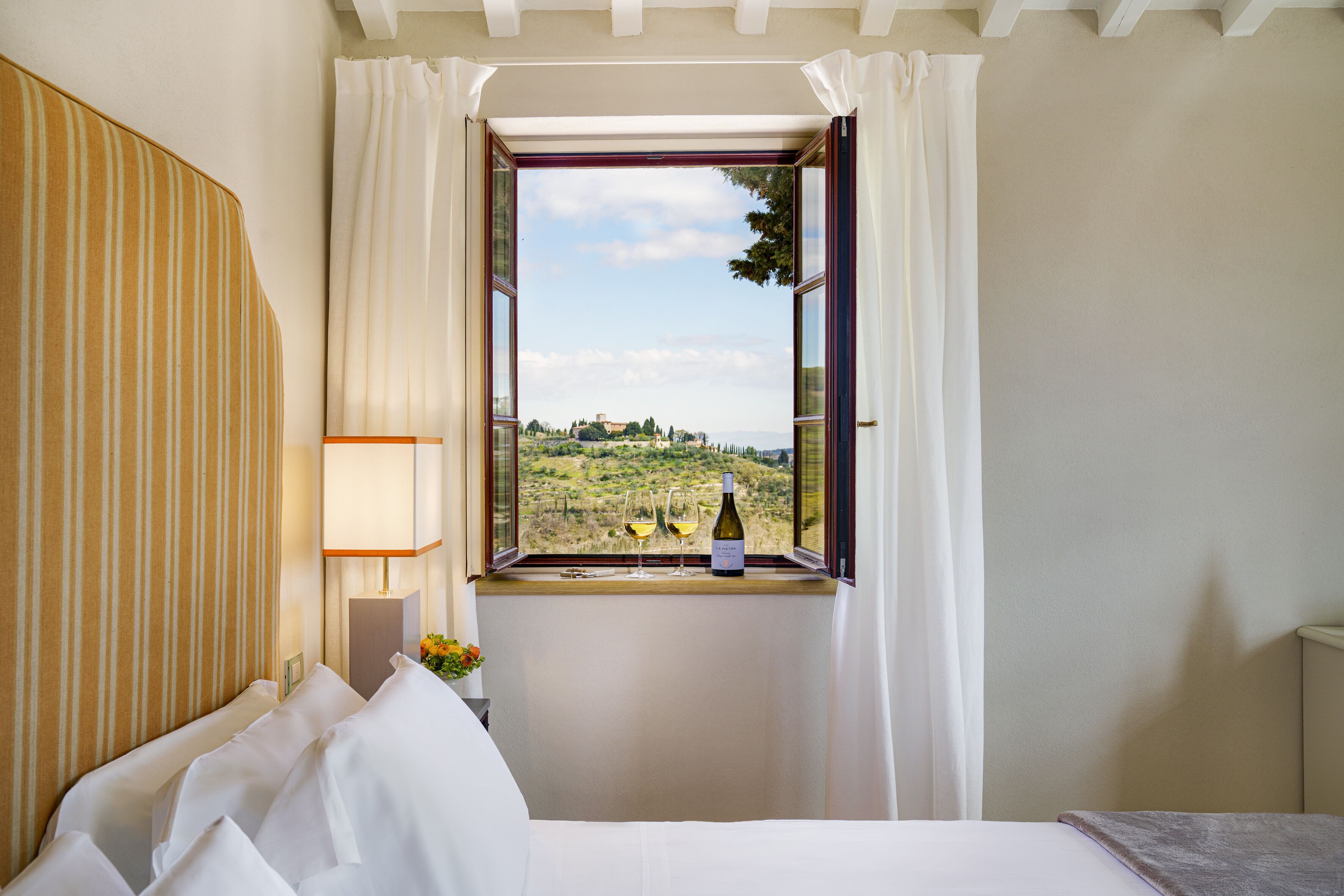 A hotel room with a bed, a window with curtains, a bedside table with a lamp, and a bottle of wine on the windowsill.