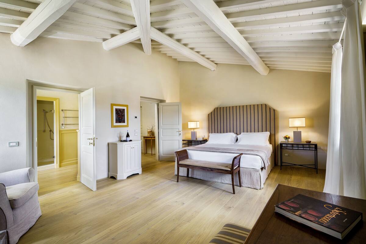 A hotel room with a wooden ceiling, a bed with white linens, a bedside table with a lamp, and a bench.
