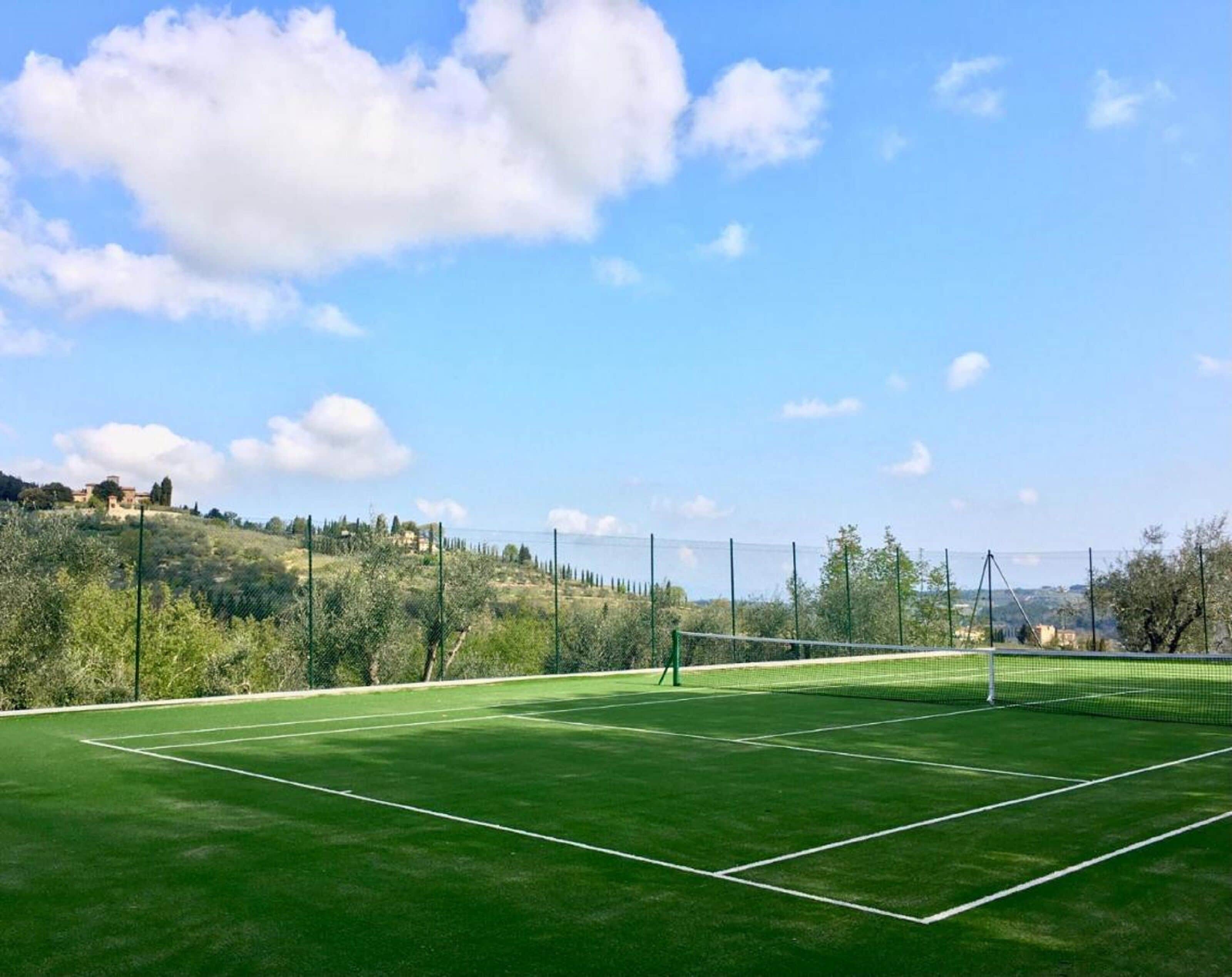 A tennis court with a net, surrounded by a fence and trees, with a clear sky above.