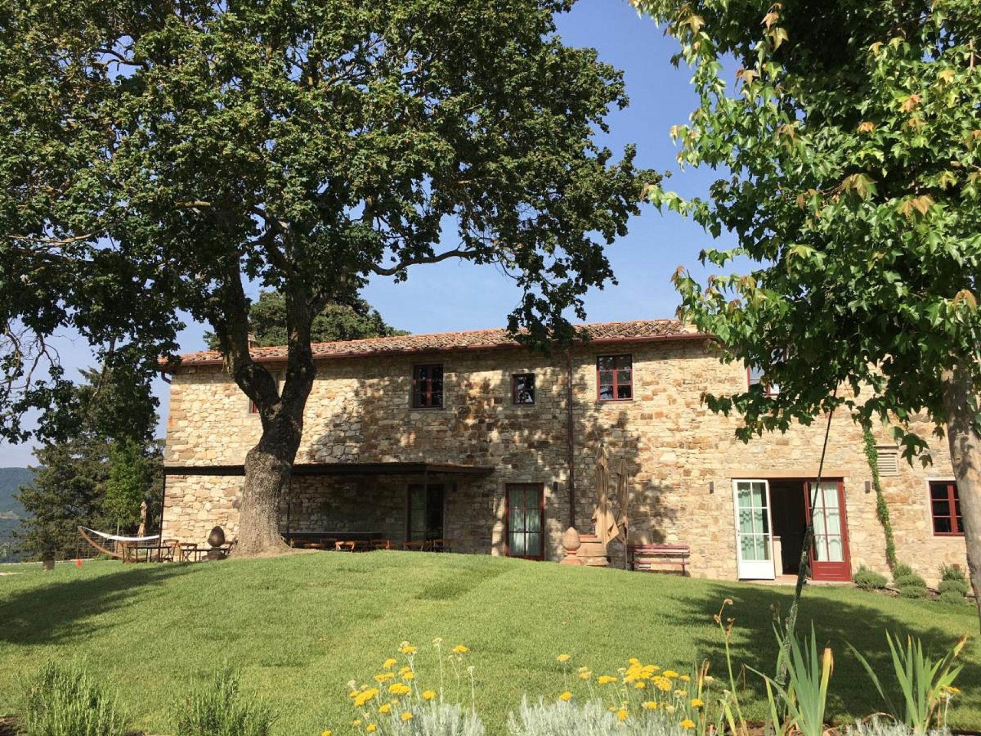 A stone building with a tiled roof, a large tree in front, and a grassy lawn with yellow flowers.