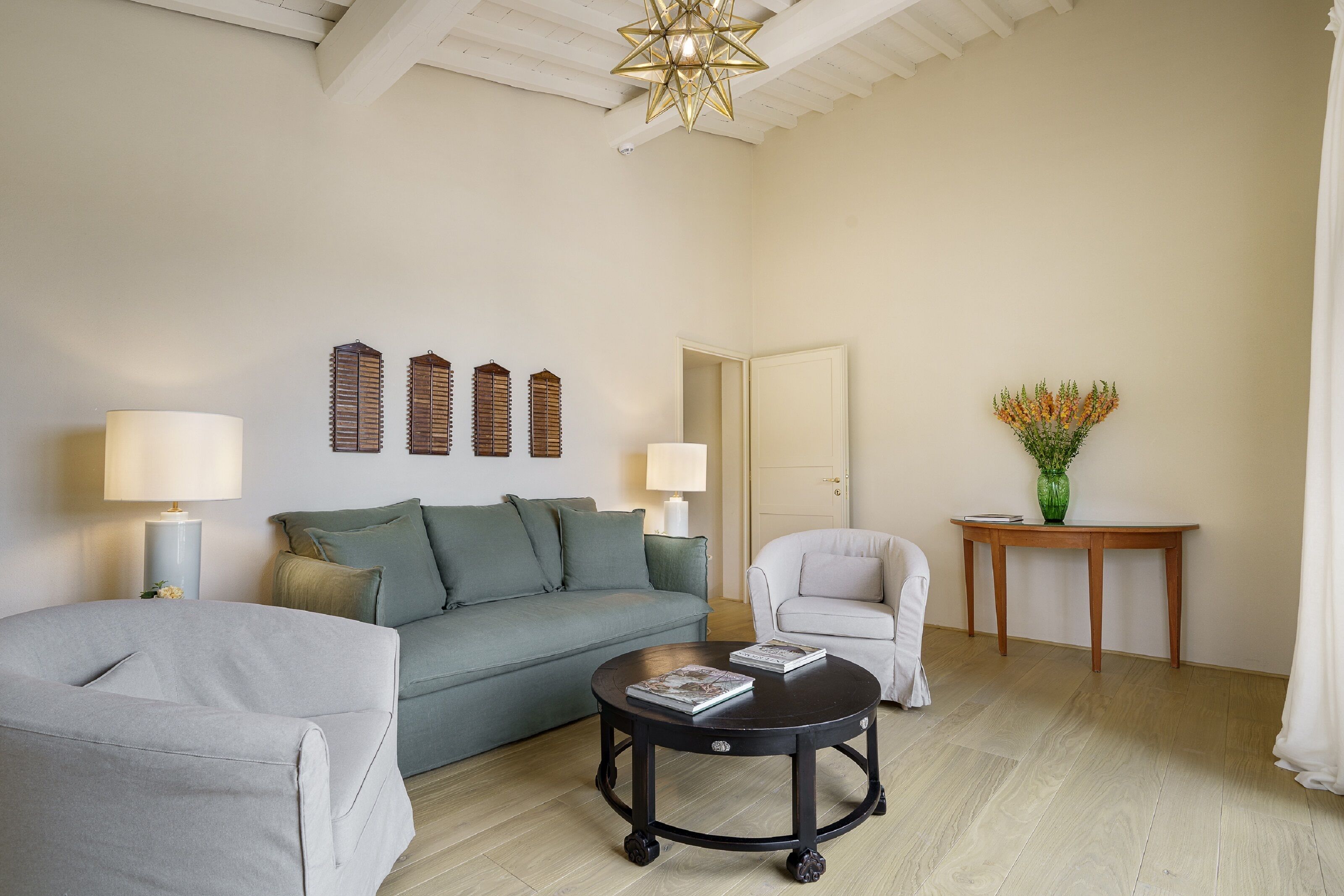 Sala de estar com sofá, poltrona, mesa de centro redonda e luminárias. Há uma porta e uma pequena mesa com um vaso.