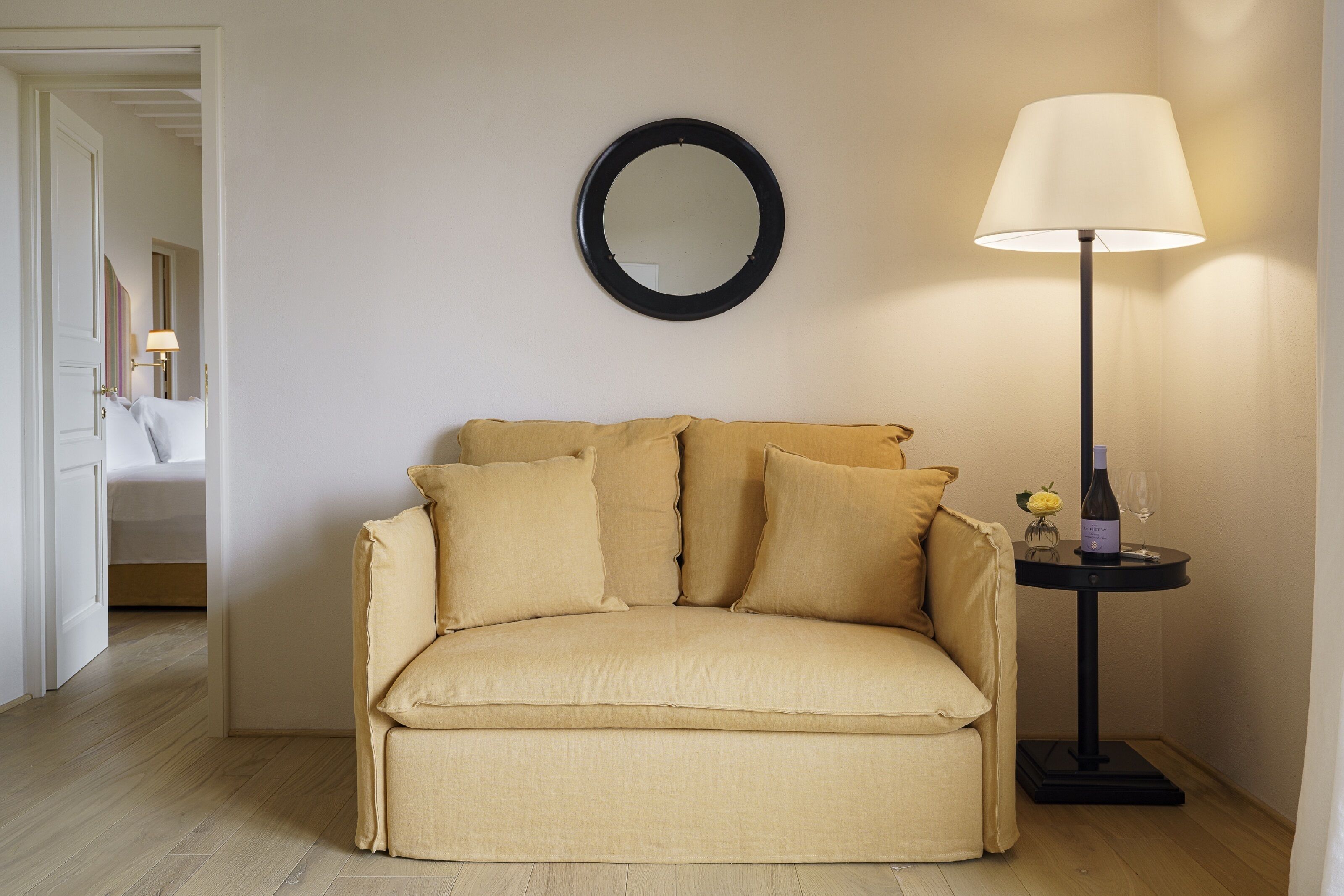 A beige sofa with multiple cushions, a round mirror on the wall, a floor lamp, and a small table with a bottle and glass.