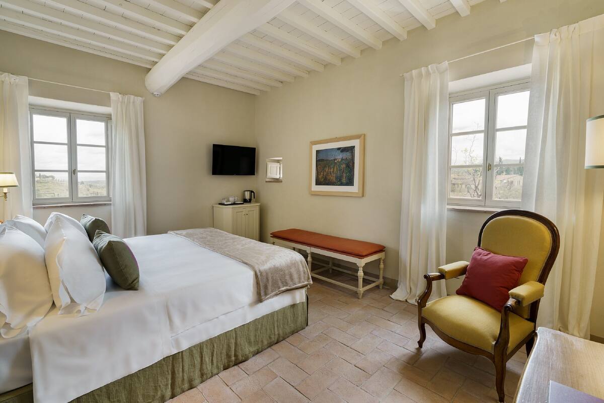 A bedroom with a bed, a chair, a bench, a television, and a painting on the wall.