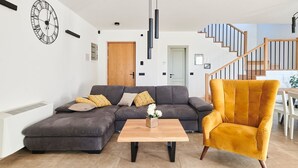 A modern living room with a grey sectional sofa, a wooden coffee table, and a mustard-colored armchair. A large wall clock and a staircase are visible in the background. - Charming Villa Grgo with a pool and a mini wellness (Tinjan)