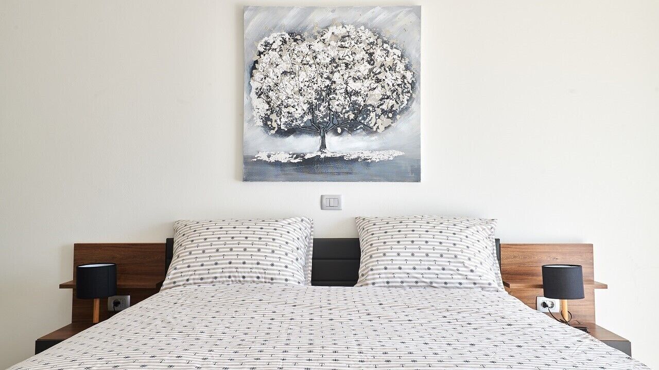 A bedroom with a bed, two nightstands, a wall-mounted lamp, and a painting of a tree.