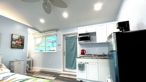 Un espacio habitable compacto con kitchenette, una cama con edredón colorido, ventilador de techo y ventana con vistas al exterior.