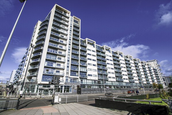 A modern multi-story residential building with balconies, a gated entrance, and a paved walkway.