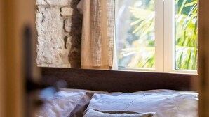 Quarto com parede de pedra, cama com roupa de cama branca e marrom, e vista para o verde pela janela.
