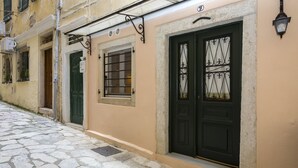 Une ruelle étroite pavée, bordée d’un immeuble dont la porte est peinte en noir et ornée de motifs en fer forgé, avec une petite fenêtre protégée par une grille métallique et un luminaire placé au-dessus de la porte.