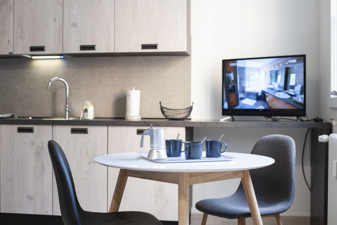 A modern kitchen with a round table, two chairs, a coffee maker, and a television displaying a room.
