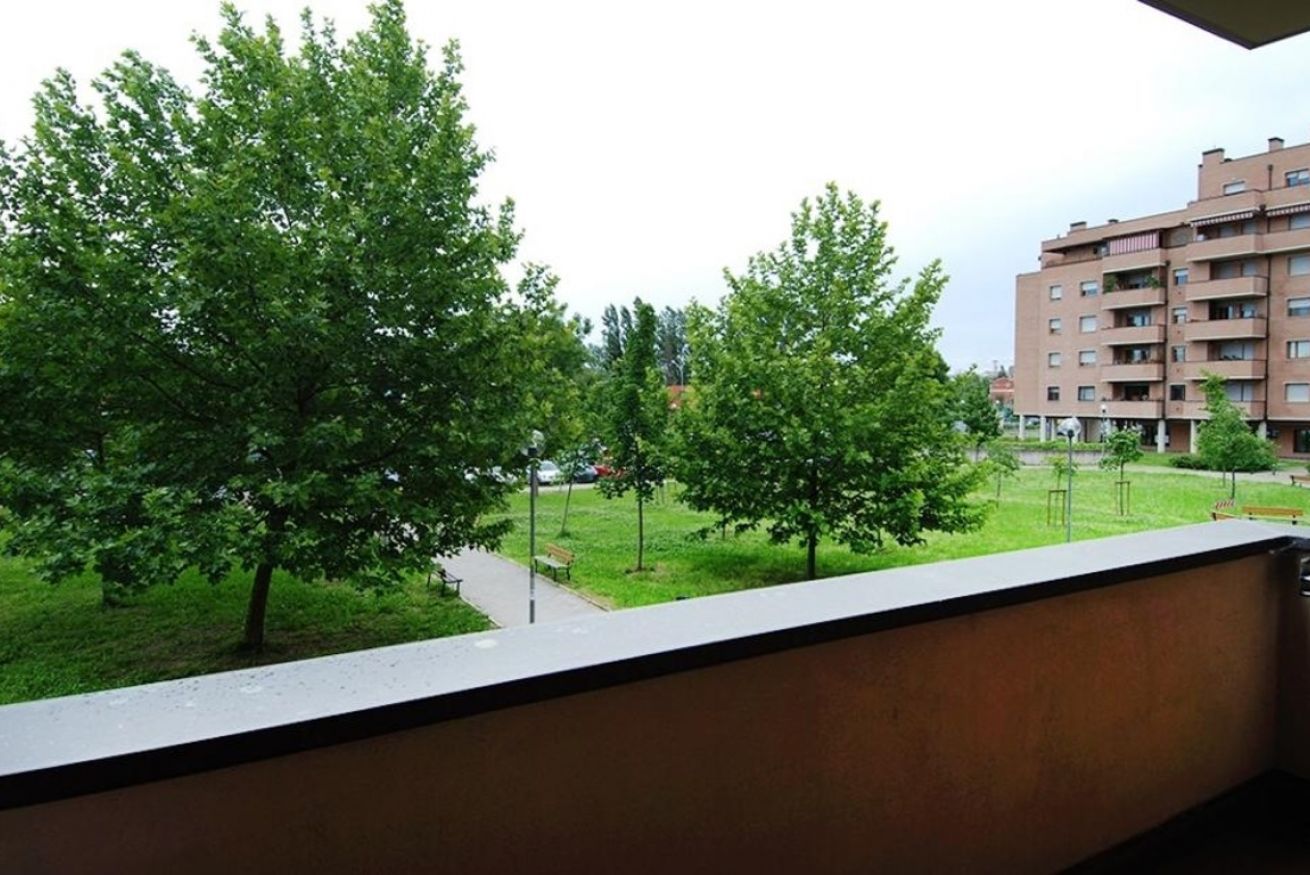 A balcony with a view of a grassy area, trees, and a multi-story building in the background.