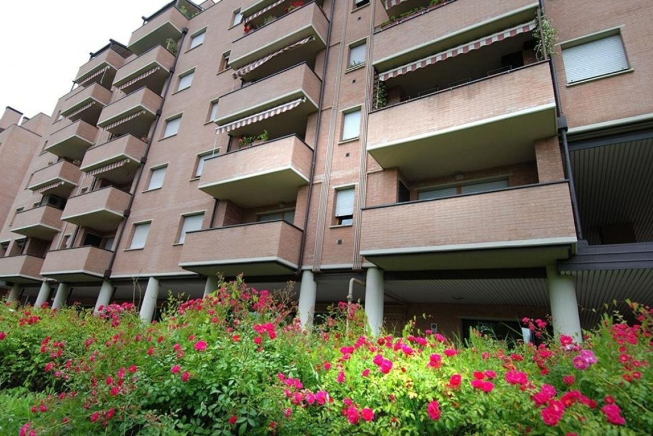 A multi-story residential building with balconies, brick exterior, and a garden with pink flowers.