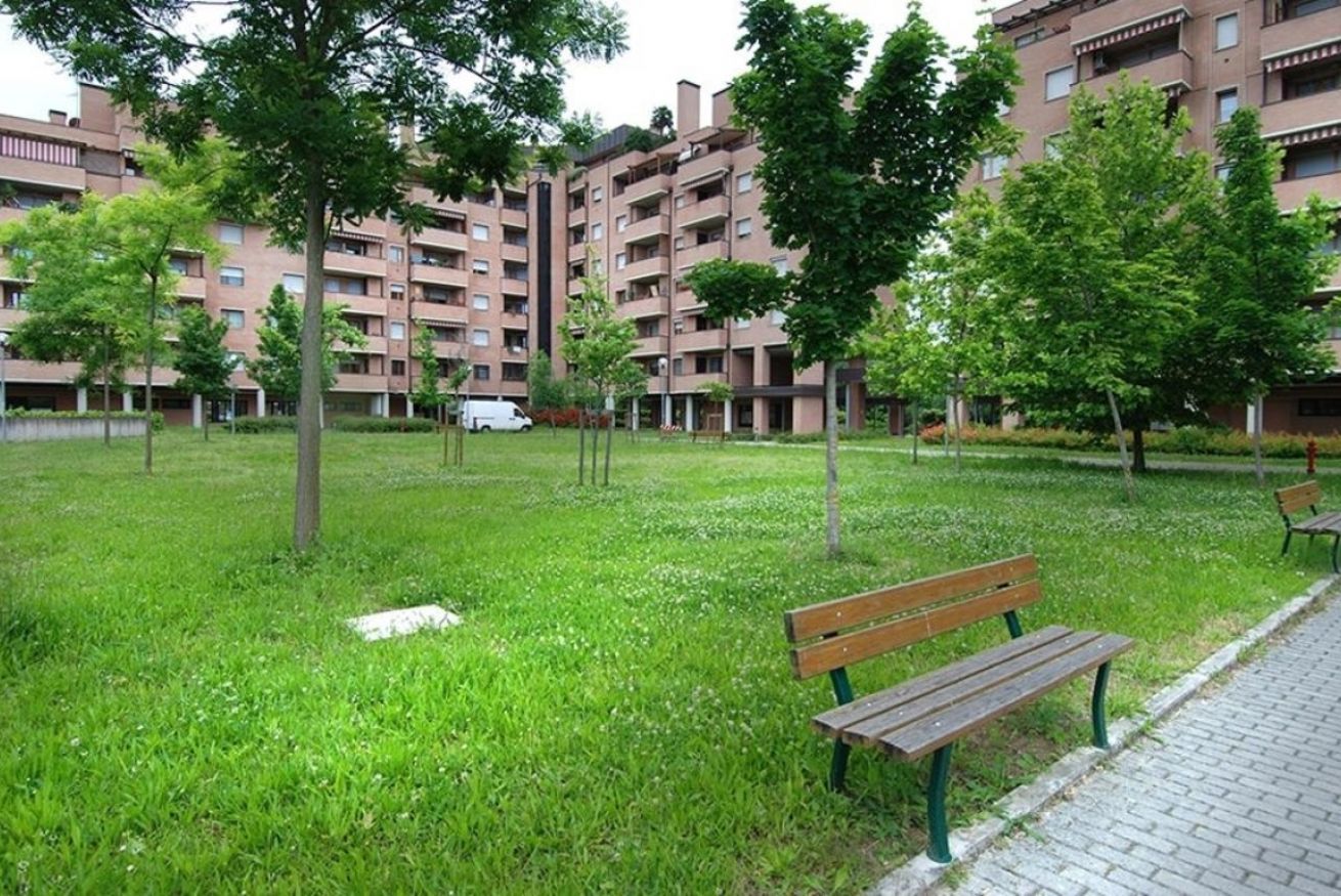 Un'area verde con prato, una panchina e alberi, circondata da edifici residenziali.