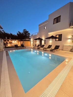 A modern outdoor swimming pool with lounge chairs and umbrellas, surrounded by a well-lit patio area.