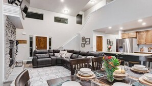 A spacious living area with a dark leather sectional sofa, a dining table set for six, and a kitchen in the background.