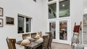 A dining area with a wooden table set for four, surrounded by wooden chairs. There is a window with a view of the outside, and a door leading to another room.