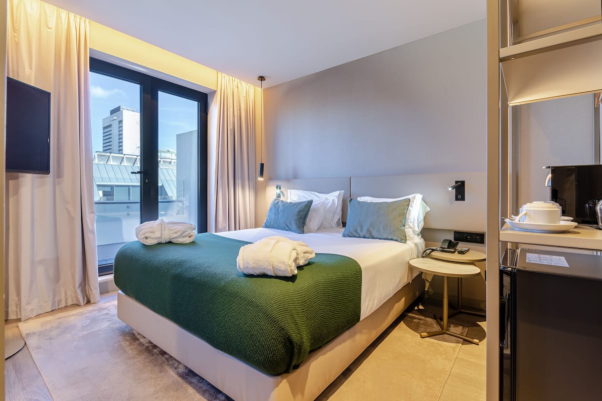 Ein Hotelzimmer mit Bett, kleinem Tisch, Kühlschrank und Blick auf die Stadt durch große Fenster.