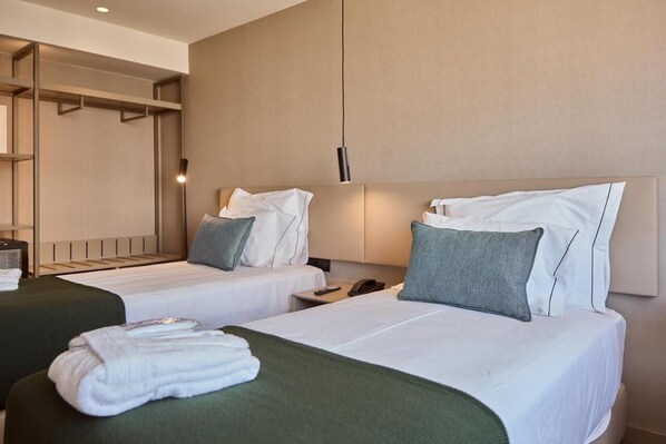 A hotel room with two beds, each with white linens and green throw pillows. There is a bedside table with a lamp, a phone, and a folded towel. The room has a neutral color scheme with beige walls and a wooden headboard.