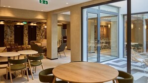 Un hall d’hôtel moderne avec des tables rondes et des chaises vertes, une porte vitrée donnant sur l’extérieur et un mur de marbre avec une porte.