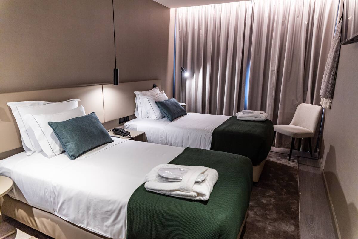 Ein Hotelzimmer mit zwei Betten, einem Sessel, einem Schreibtisch und einem großen Fenster mit Vorhängen.
