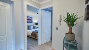 A hallway with a bed in the background, a potted plant on a stand, and a door leading to another room.