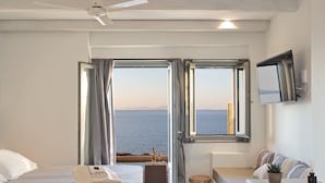 Habitación de hotel con cama, una pequeña mesa con una botella y vasos, zona de estar con una planta y vista al mar.