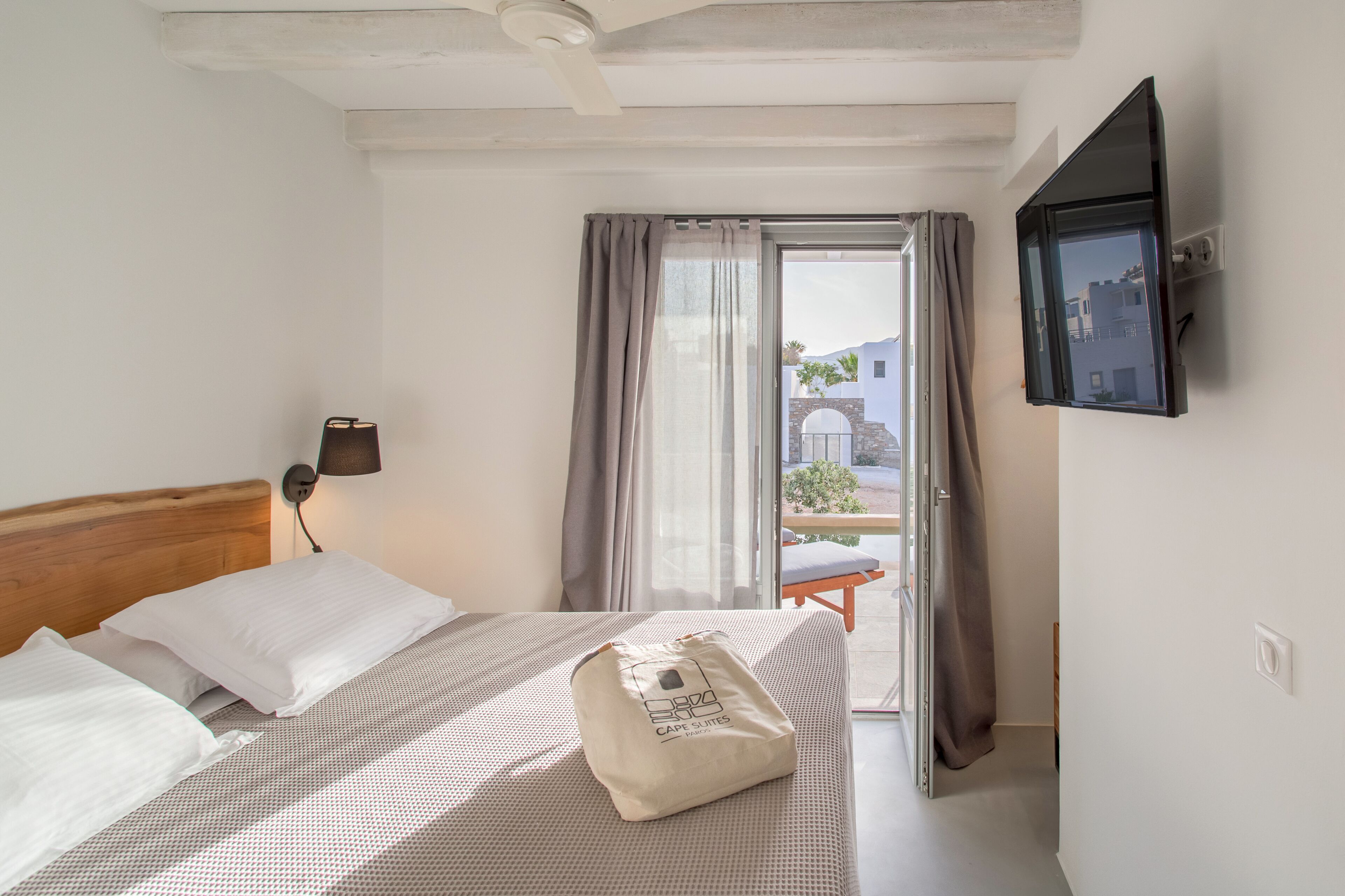 A hotel room with a bed, a bedside lamp, a TV mounted on the wall, and a view of the outside through a window with curtains.