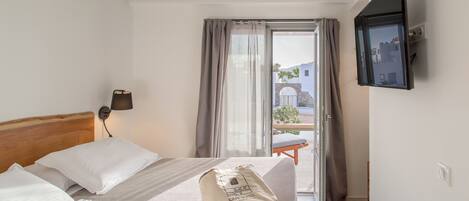 A hotel room with a bed, a bedside lamp, a TV mounted on the wall, and a view of the outside through a window with curtains.
