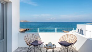 Un balcón con dos sillones de mimbre y una mesita pequeña, con vistas a un mar azul cristalino y una costa rocosa.