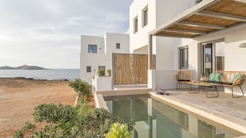 Uma área de piscina moderna ao ar livre, com vista para o mar, uma pérgola de madeira e assentos com almofadas.