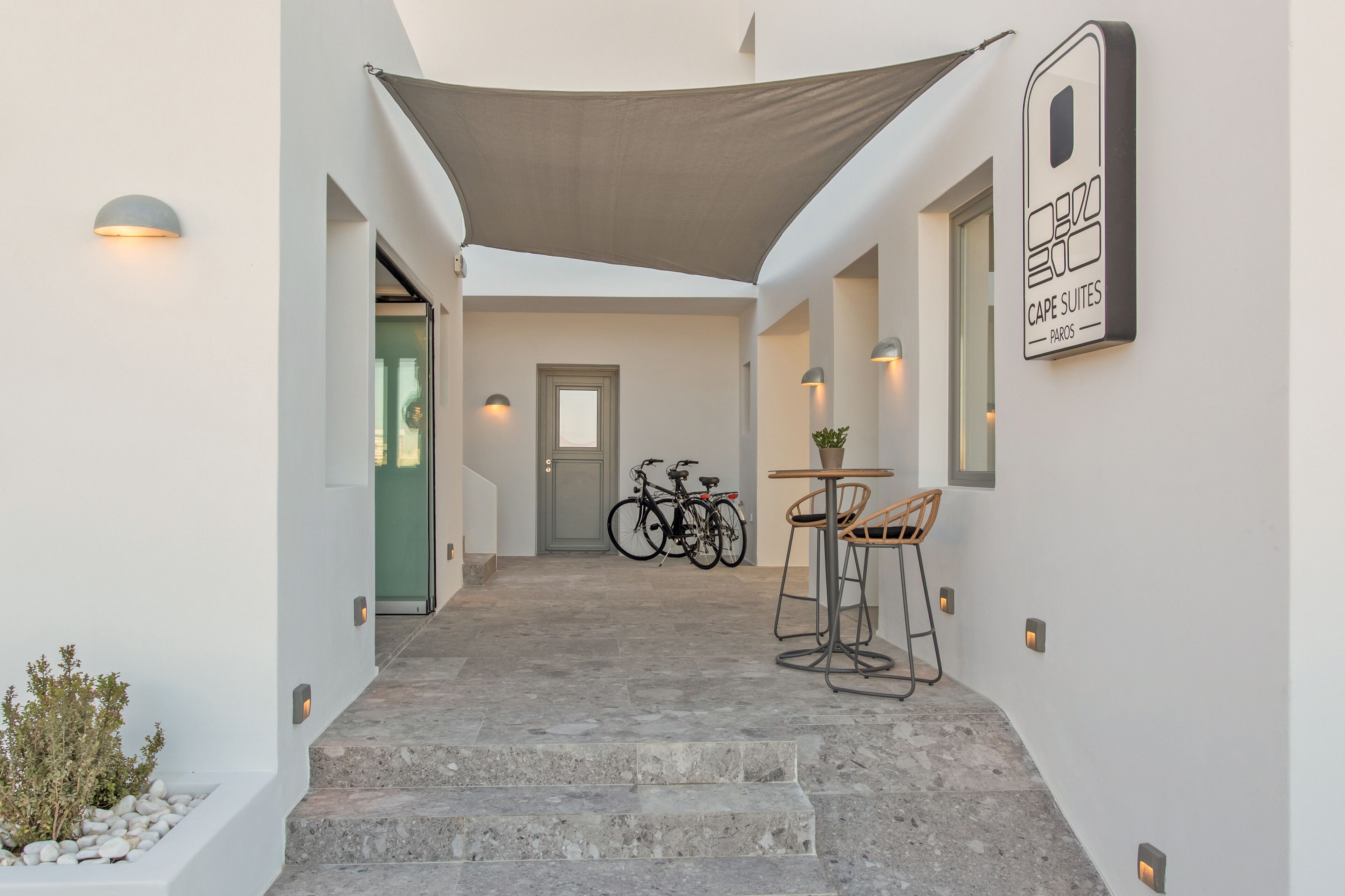 A modern hotel corridor with a covered outdoor seating area, a bicycle, and a sign for Cape Suites Paros.