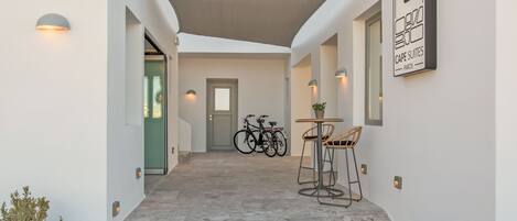 A modern hotel corridor with a covered outdoor seating area, a bicycle, and a sign for Cape Suites Paros.