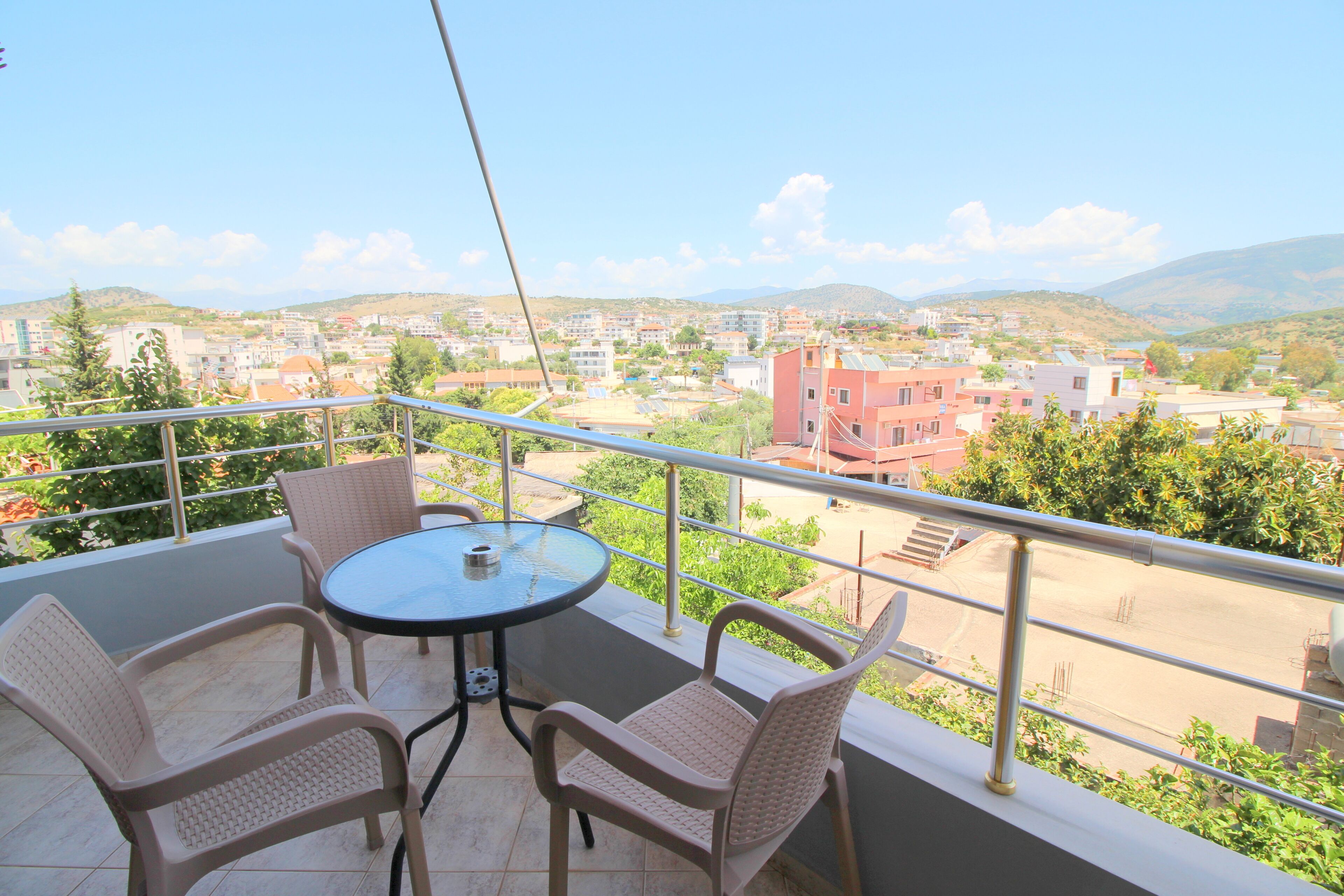 A balcony with a round table and four wicker chairs, overlooking a cityscape with buildings and hills.