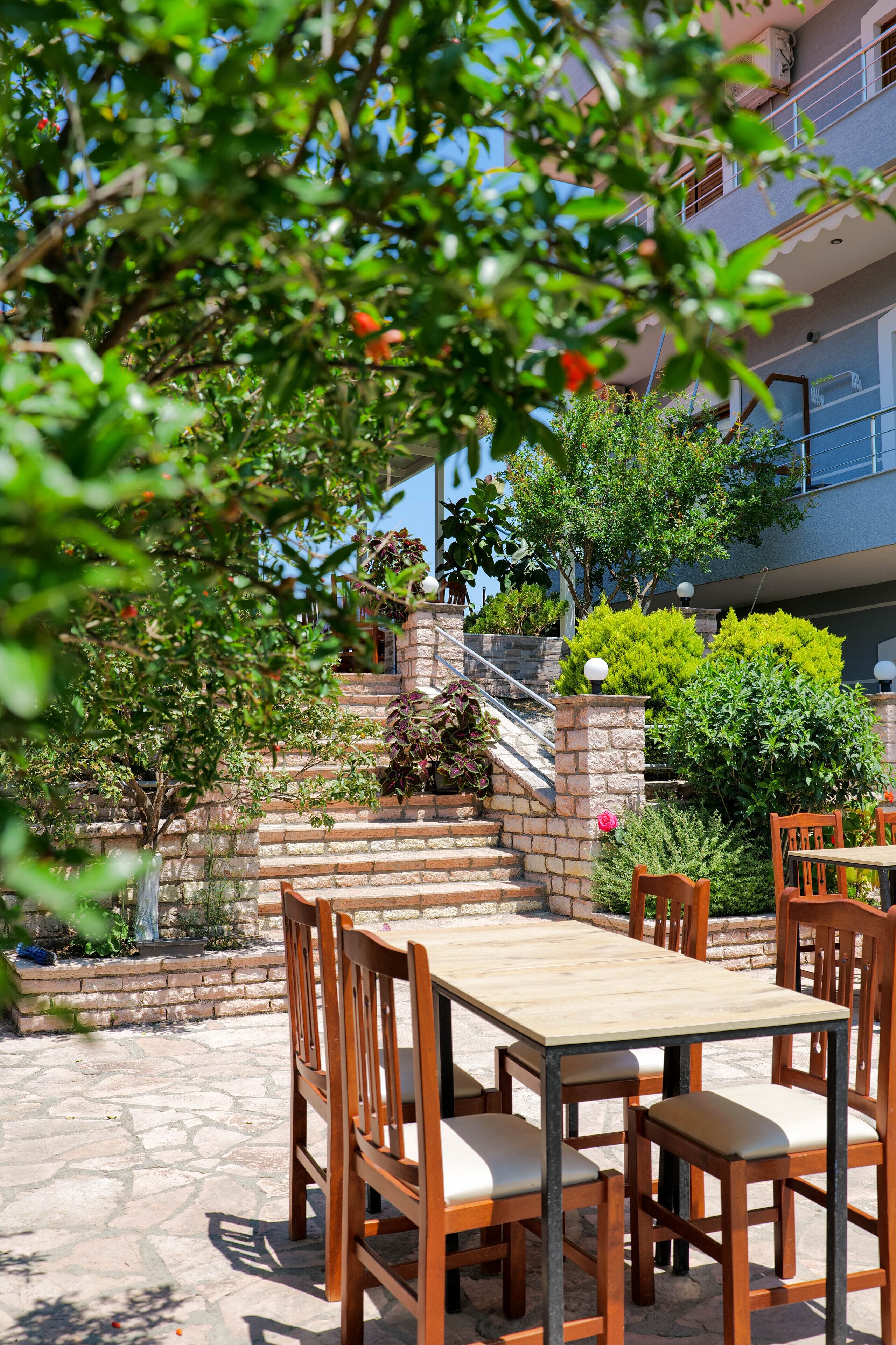 An outdoor patio with a stone floor, wooden tables and chairs, and a staircase leading to a building.