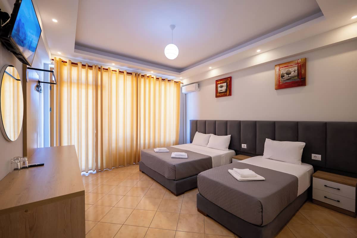A hotel room with two beds, a desk, a TV, and a large window with curtains.