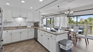 A modern kitchen with white cabinets, granite countertops, and a large island. There is a dining area with white chairs and a table, and a view of palm trees through large windows. - Gorgeous 3 bedroom 2 bath unit. Beachfront property! (Destin)